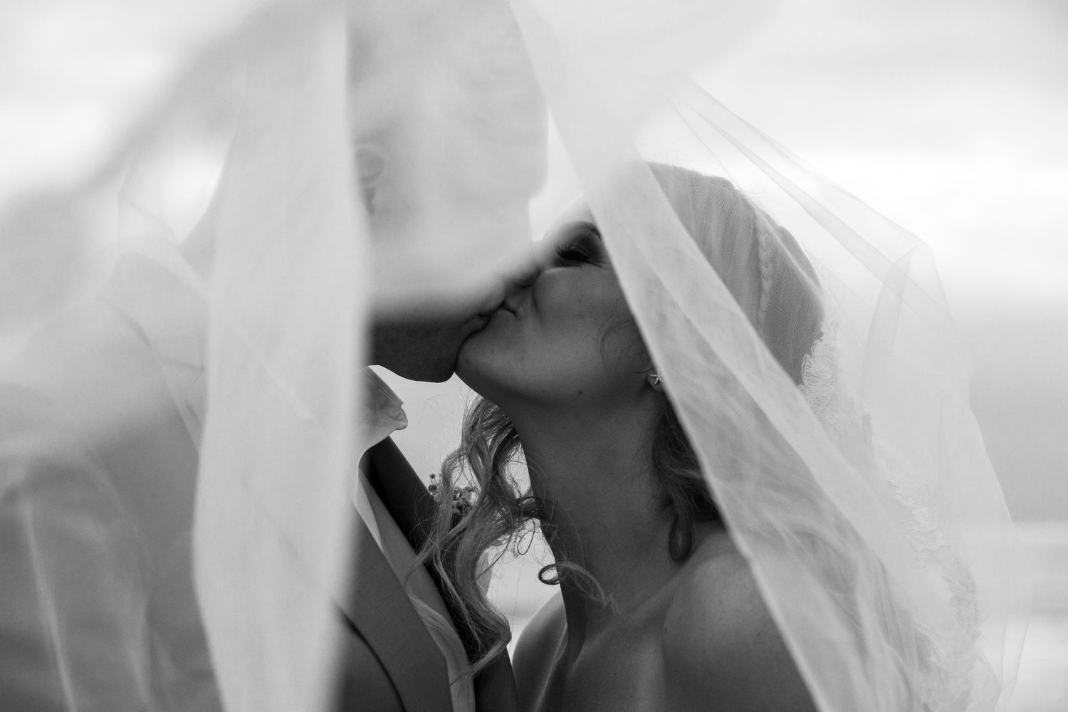 A tender wedding moment captured in black and white as a couple shares a kiss beneath a flowing veil.