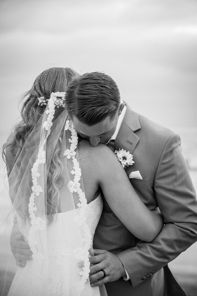 An intimate black and white wedding photo shows a tender moment between a bride in a lace veil and groom in a gray suit.