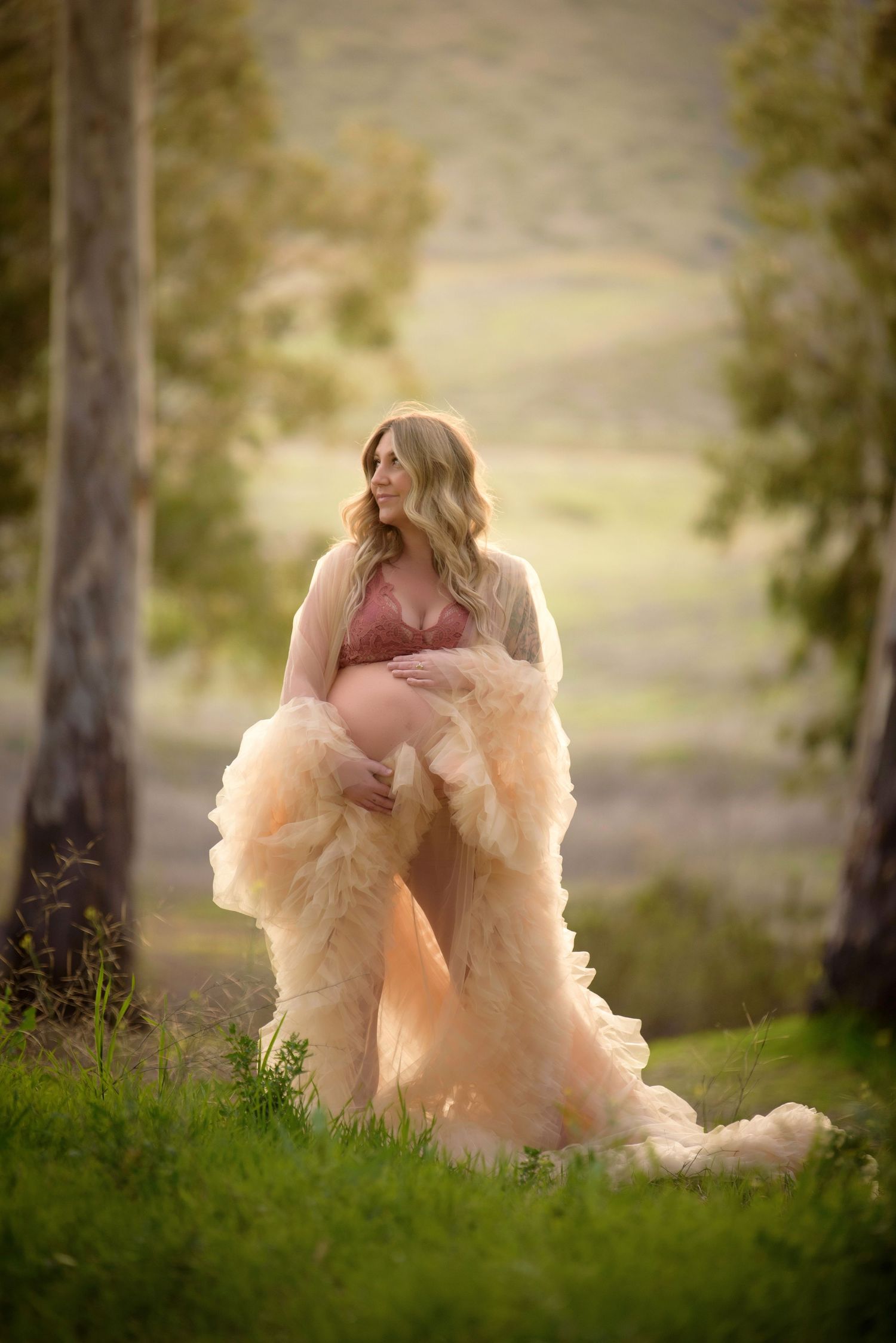 An expectant mother in pink dress and flowing peach tulle robe poses outdoors in a dreamy nature setting.