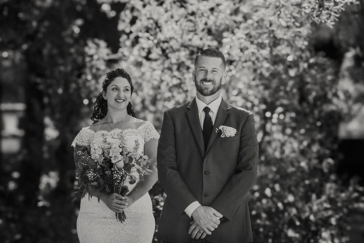 Black and white wedding photo of bride and groom standing together outdoors with spring blossoms in background.