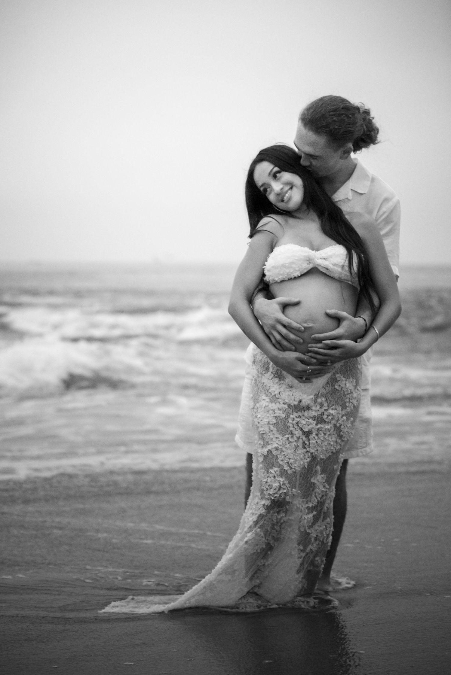 Romantic maternity photo with couple embracing on beach in black and white as waves crash behind them.