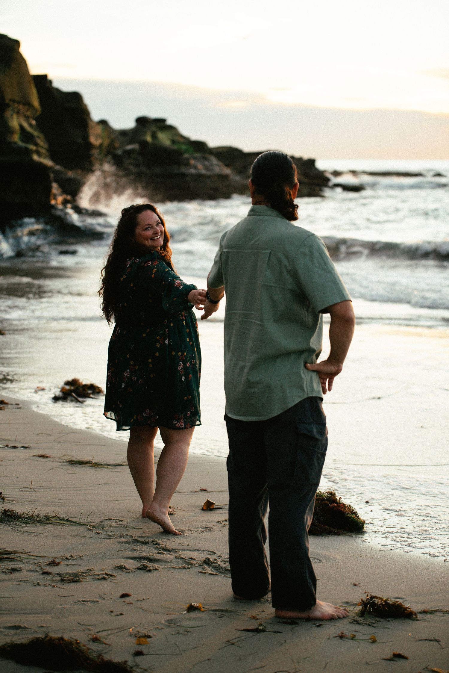 Couple dancing barefoot on a sandy beach at sunset, woman in floral dress smiling, waves crashing on rocks behind them.