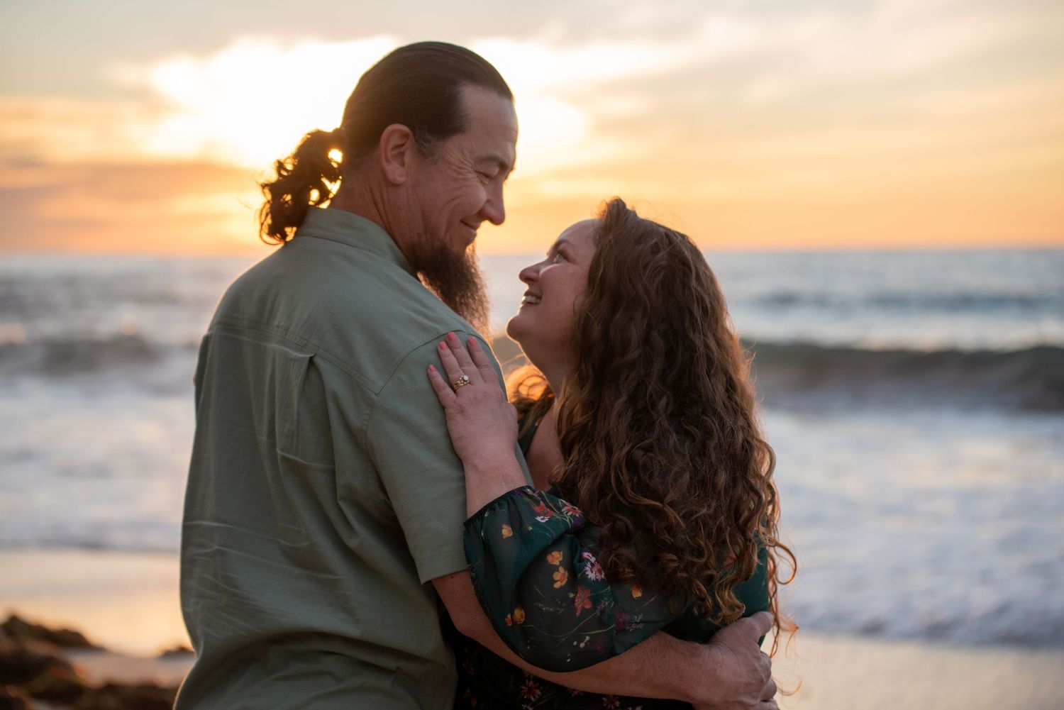 Couple embracing and smiling at each other on a beach at sunset with ocean waves in the background.