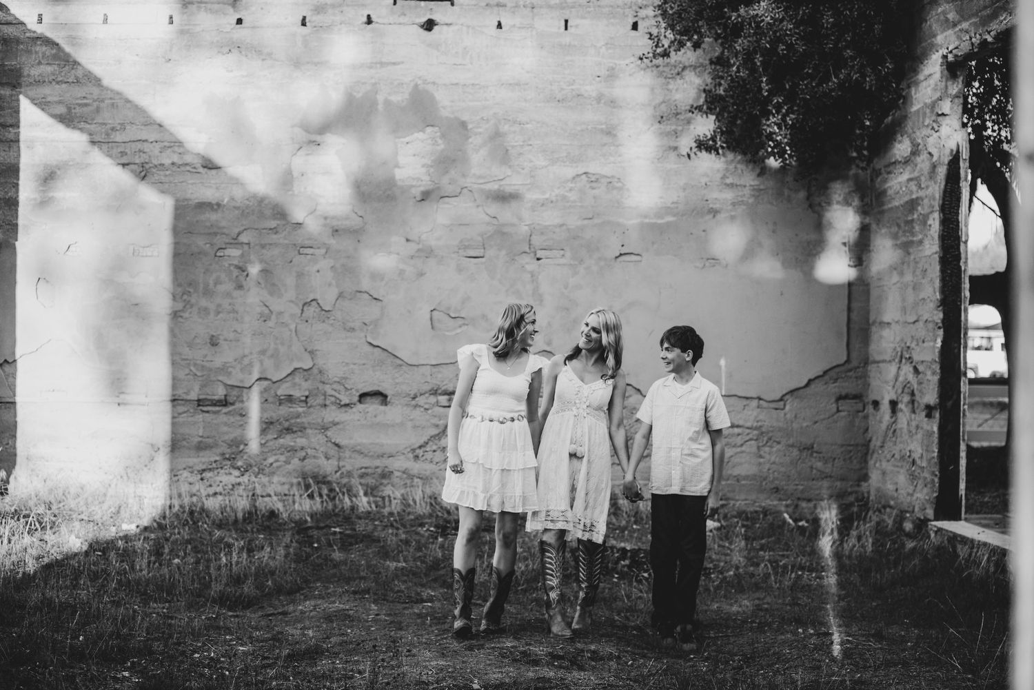 Black and white photo of three children holding hands walking near a rustic stone wall outdoors.