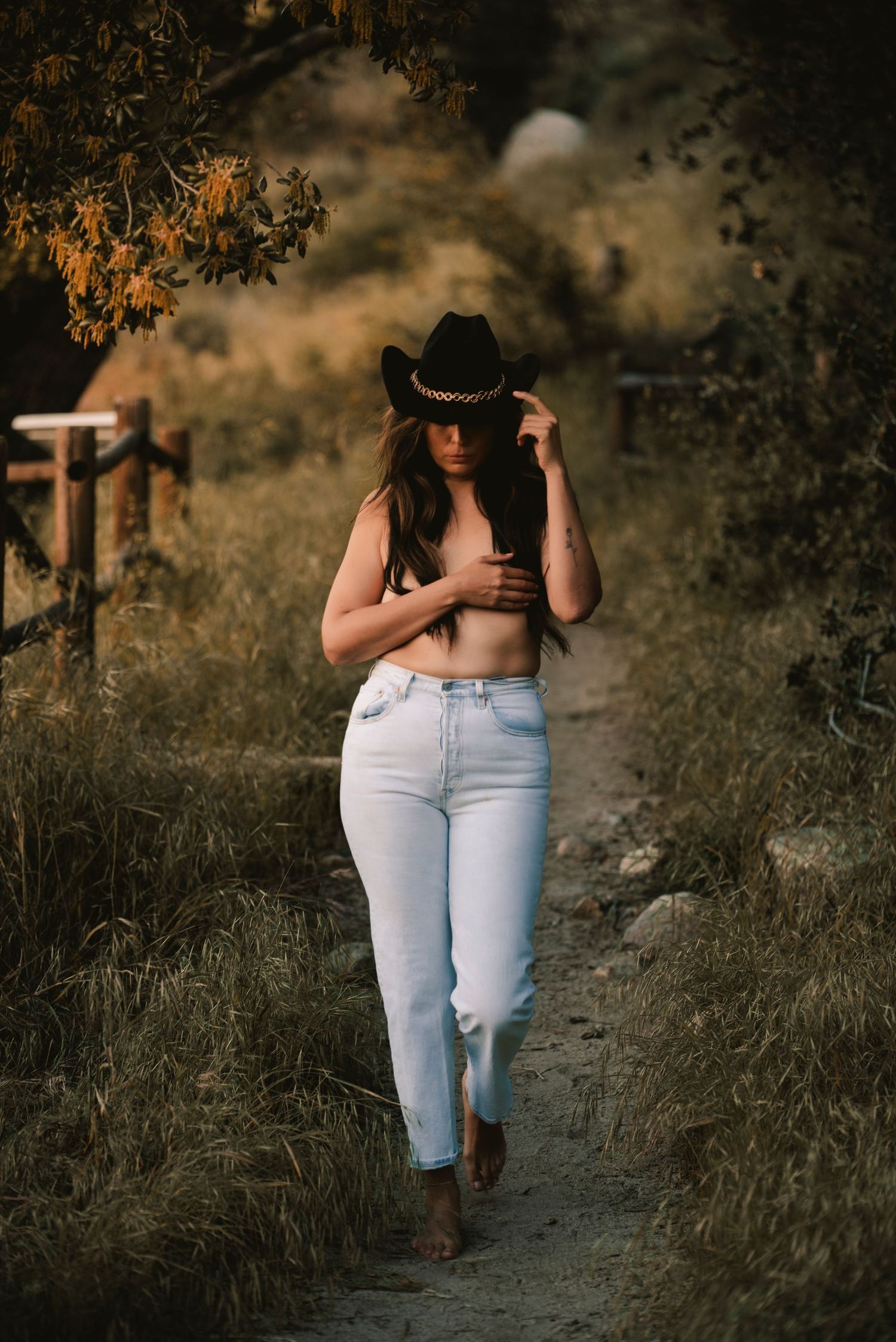 Artistic countryside portrait showing person in black cowboy hat and light denim jeans walking on rural path at sunset.