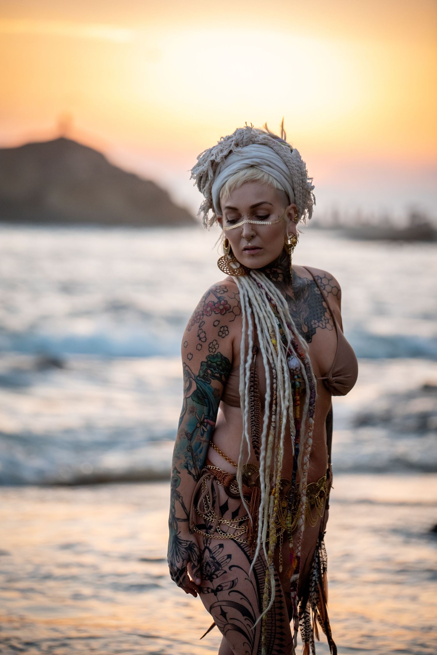 Body painted figure with white dreadlocks poses on beach at sunset with ocean waves and silhouetted mountain behind.