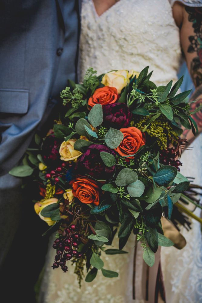 A vibrant wedding bouquet featuring orange roses, burgundy florals, yellow blooms and fresh eucalyptus greenery.