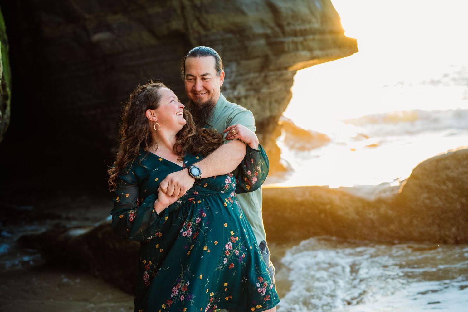 Couple laughing and dancing together on a rocky beach at golden hour, woman in floral dress, man in teal shirt.