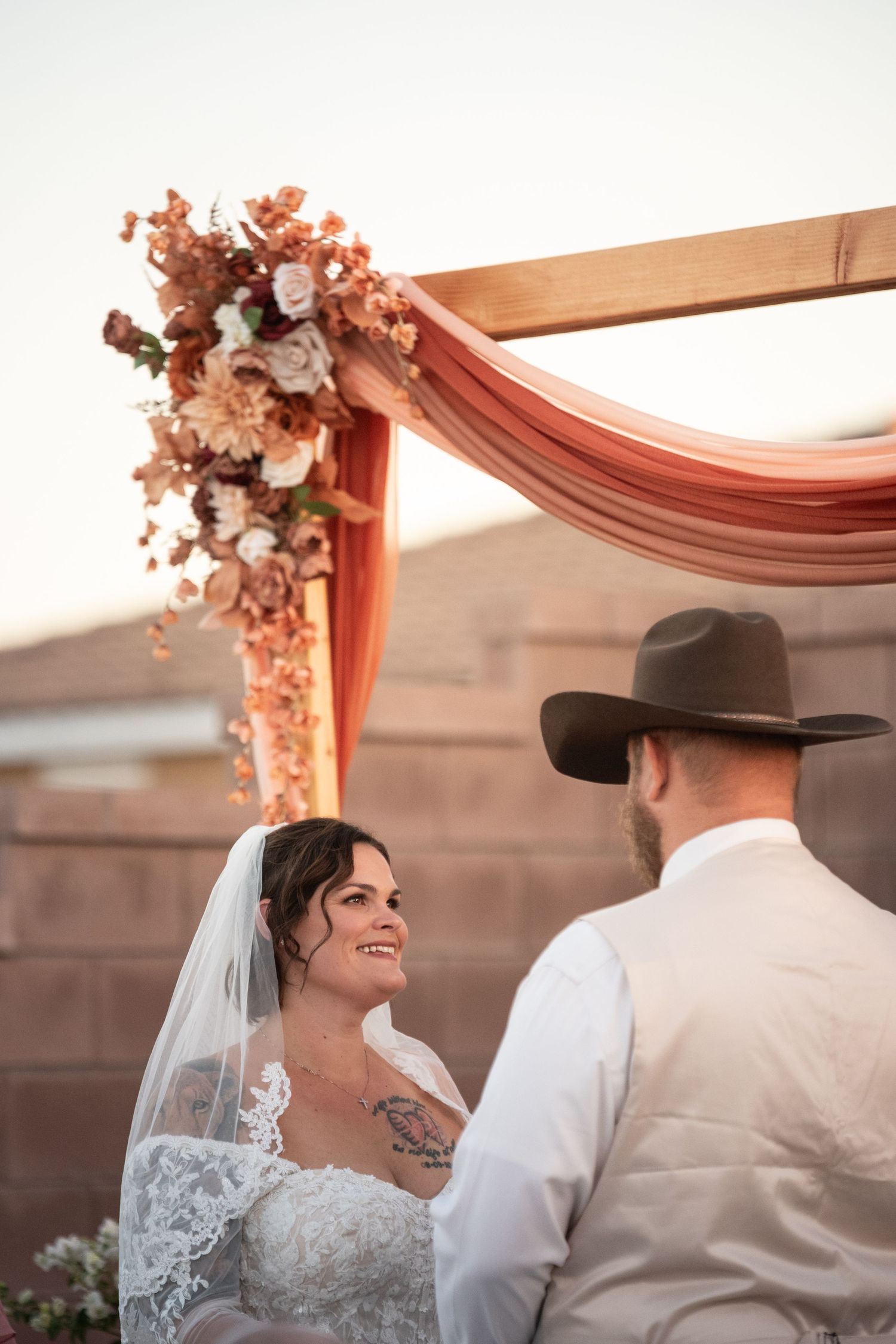Bride smiling at groom in cowboy hat during outdoor wedding ceremony under floral arch with terracotta draping.