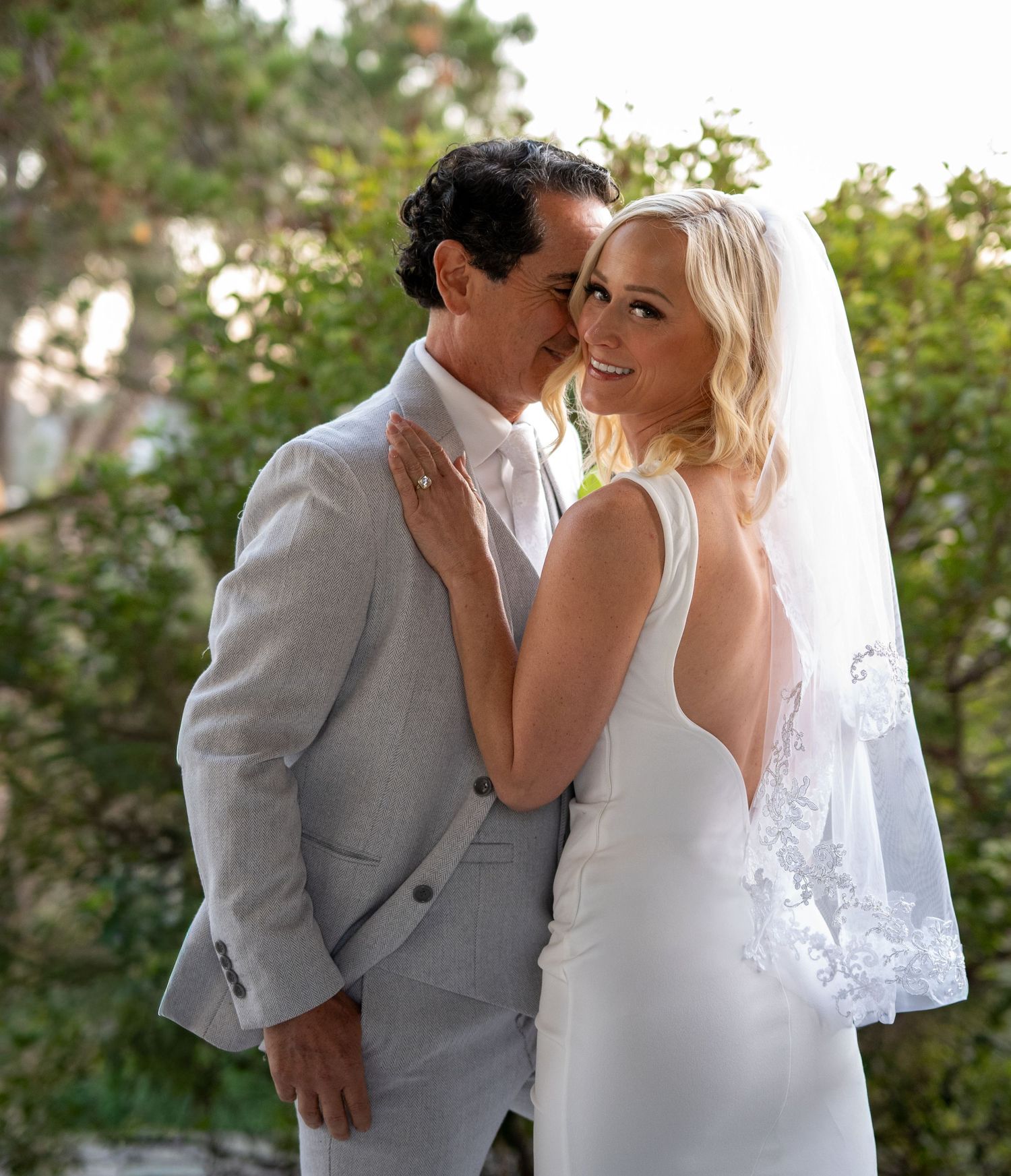 Bride in backless white gown with veil and groom in gray suit share romantic moment surrounded by greenery.