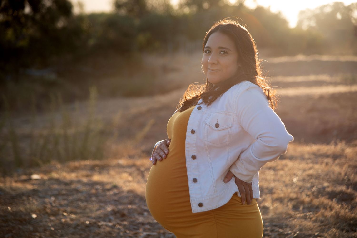 Expectant parent poses in white denim jacket and yellow dress at sunset in golden field.