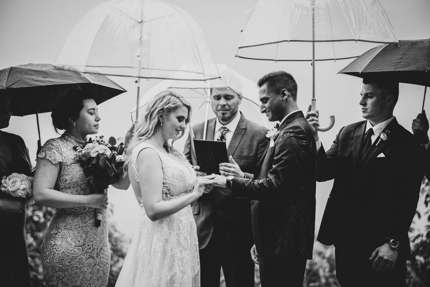 Black and white photo of people holding umbrellas during an outdoor wedding ceremony in the rain.