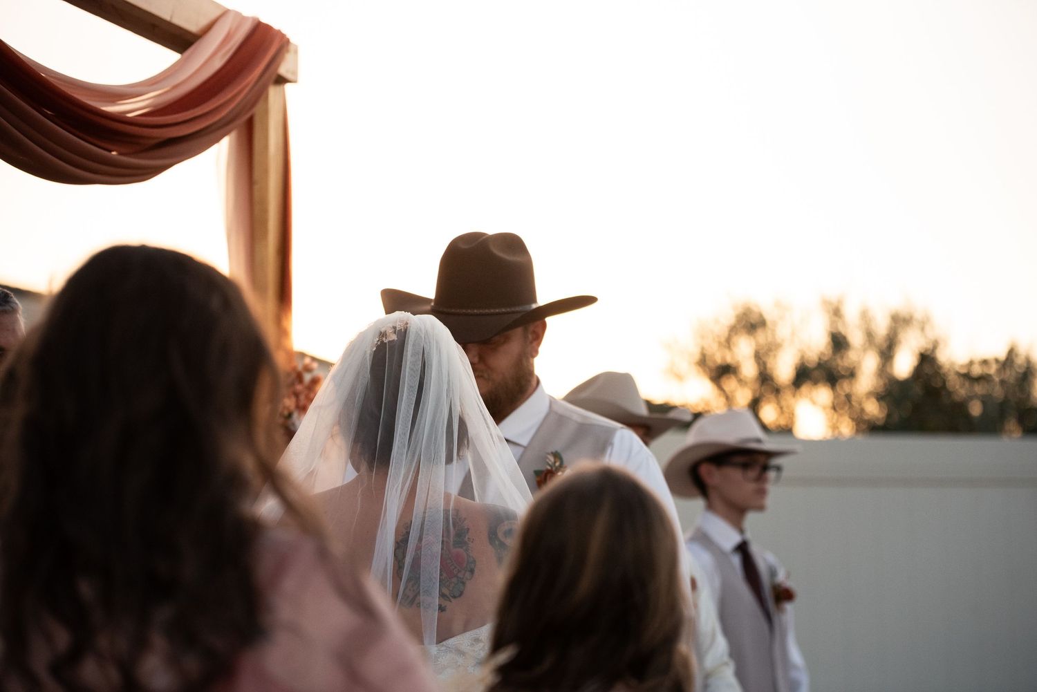 Outdoor wedding ceremony at sunset with groom in brown cowboy hat and bride in white veil standing at decorated altar.