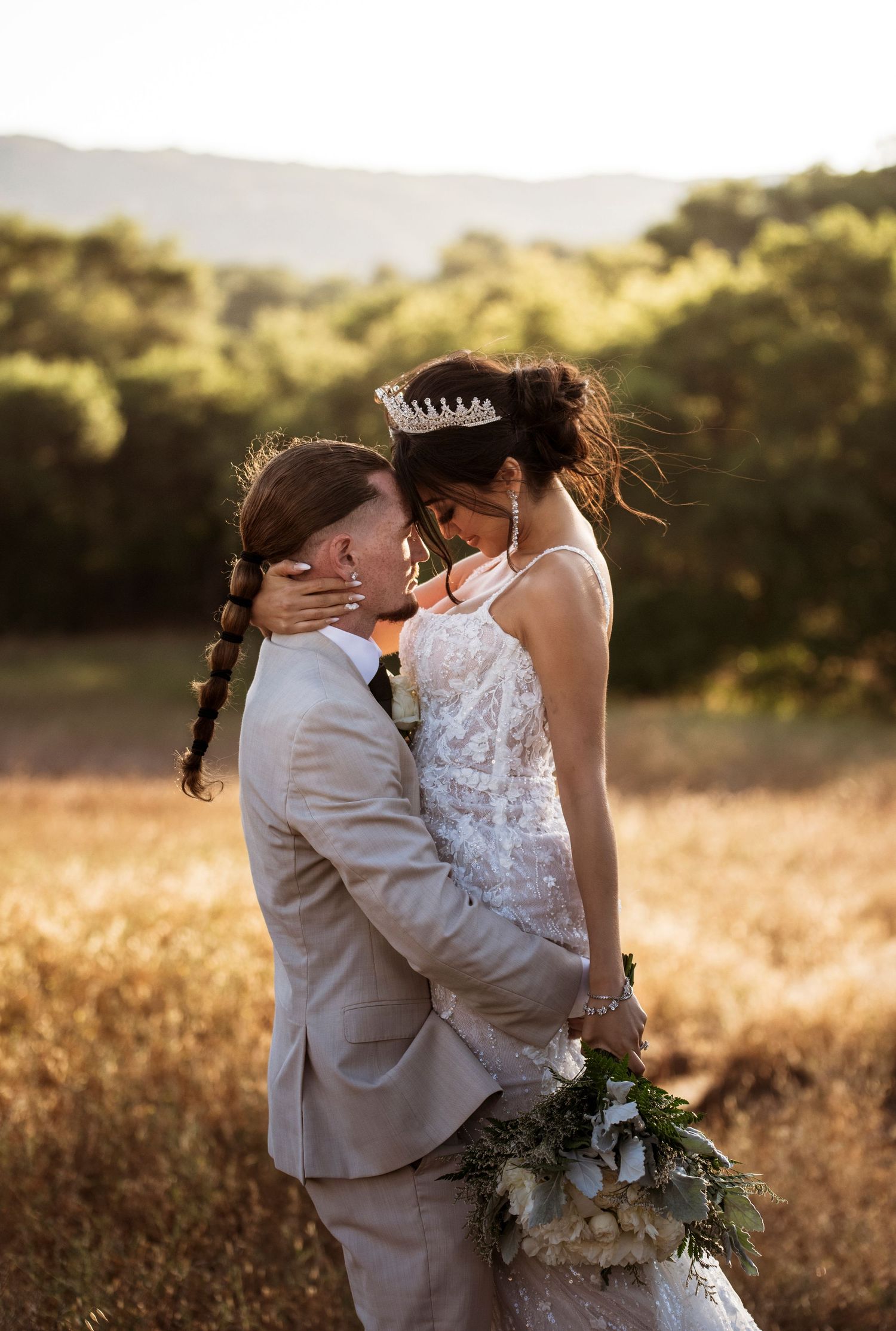 Romantic newlyweds embrace in a golden field at sunset while bride holds a rustic greenery bouquet.