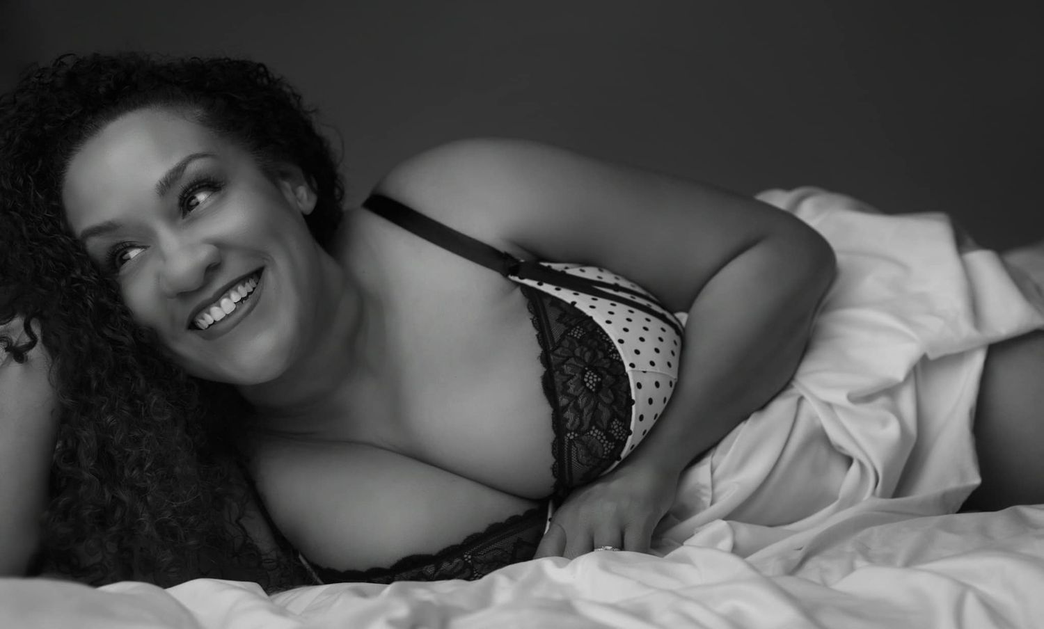 Black and white boudoir photo showing someone lying on white bedding in lingerie while smiling at the camera.