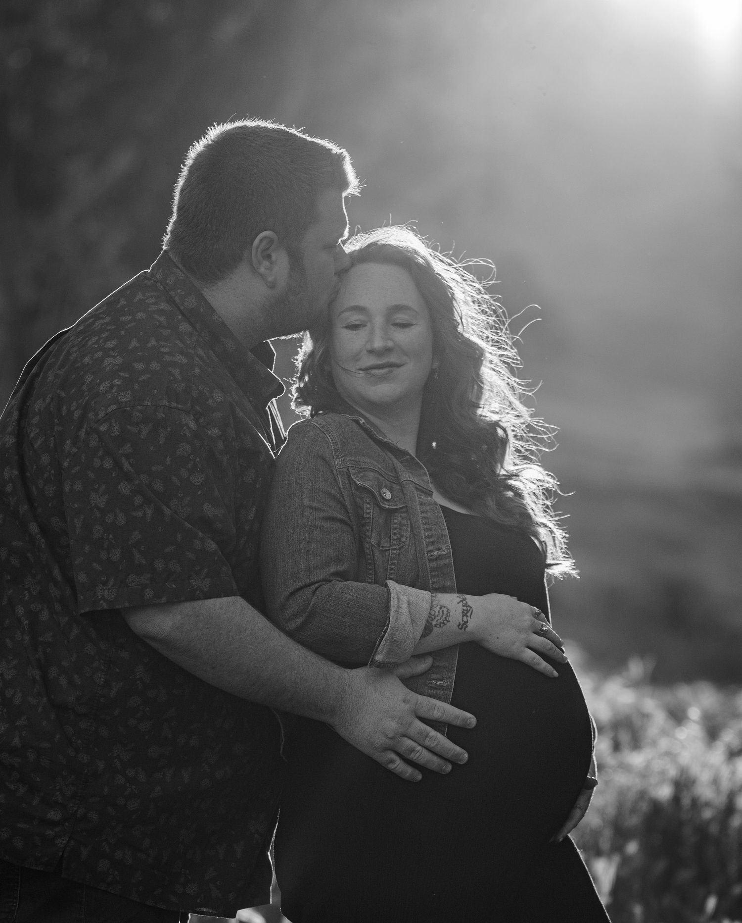 Black and white maternity photo of a couple embracing in soft backlit sunlight during an outdoor photoshoot.