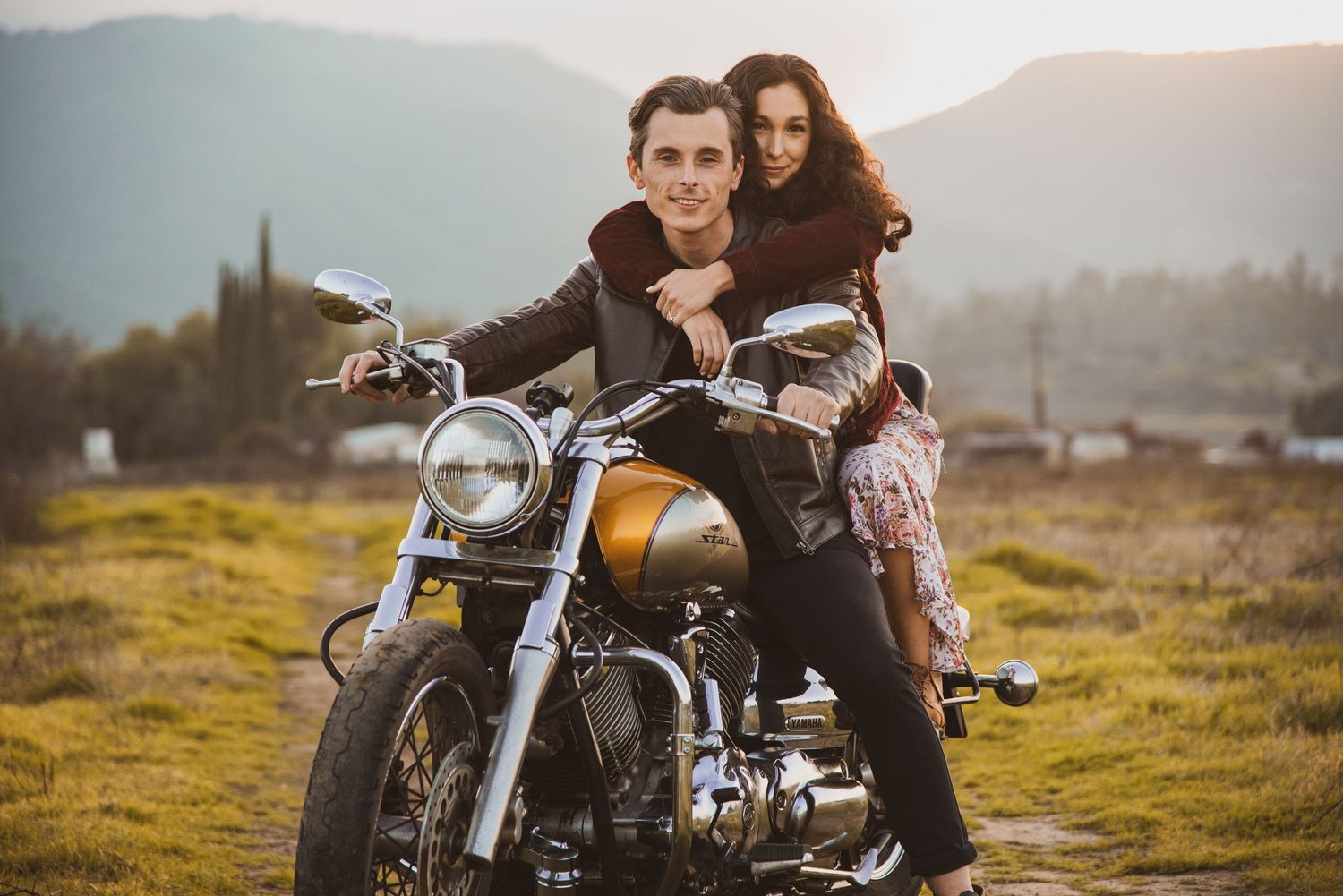 Two people ride together on a vintage motorcycle in a scenic mountain landscape at sunset.