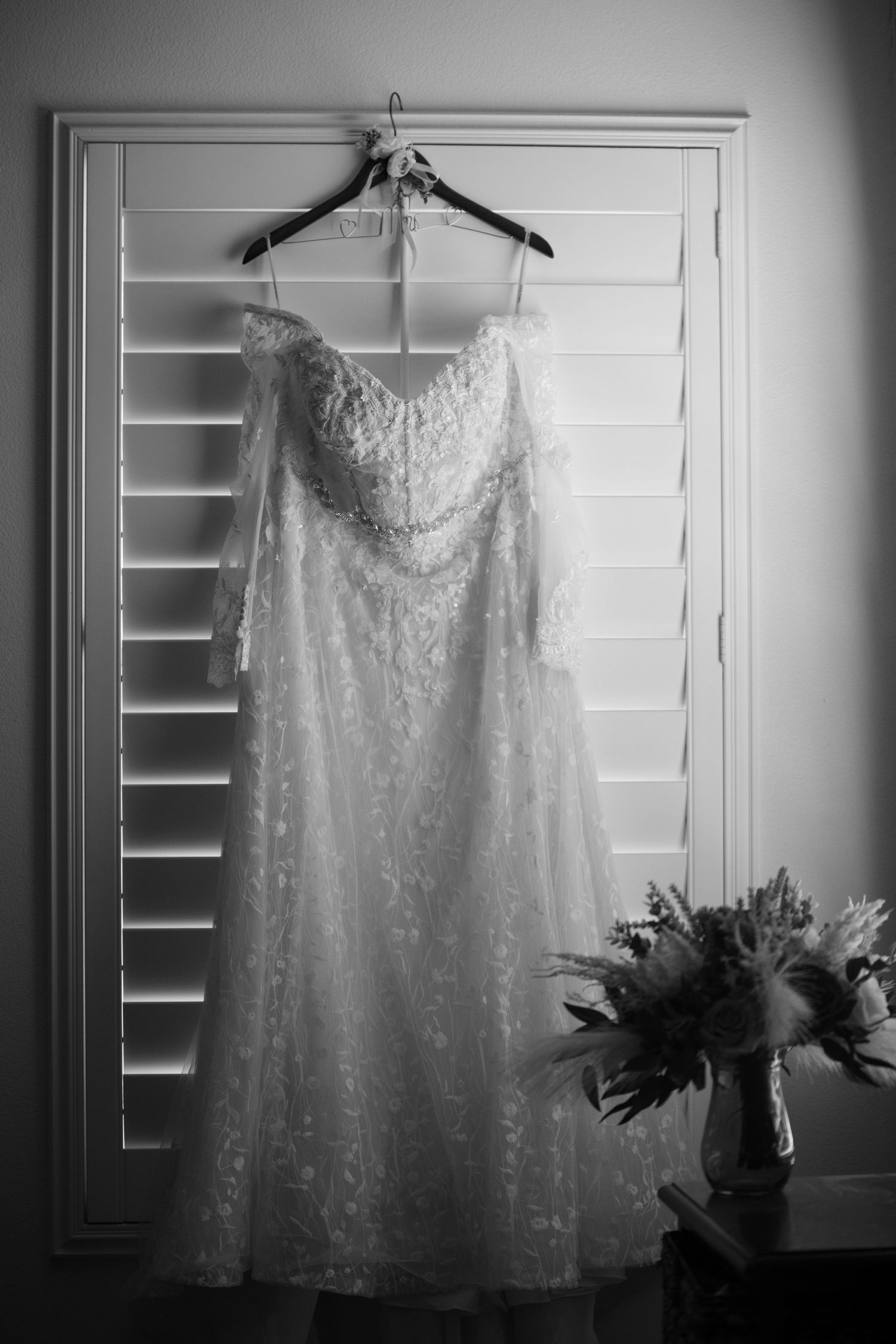 Black and white photo of an off-shoulder lace wedding dress hanging on a hanger before a shuttered window.