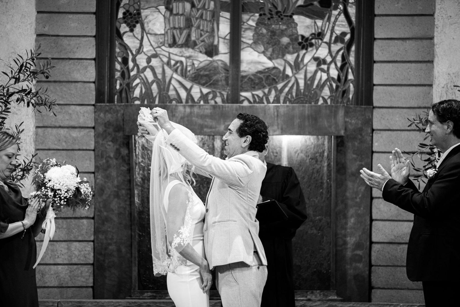 Groom lifts bride during wedding ceremony kiss in black and white photo with guests applauding.