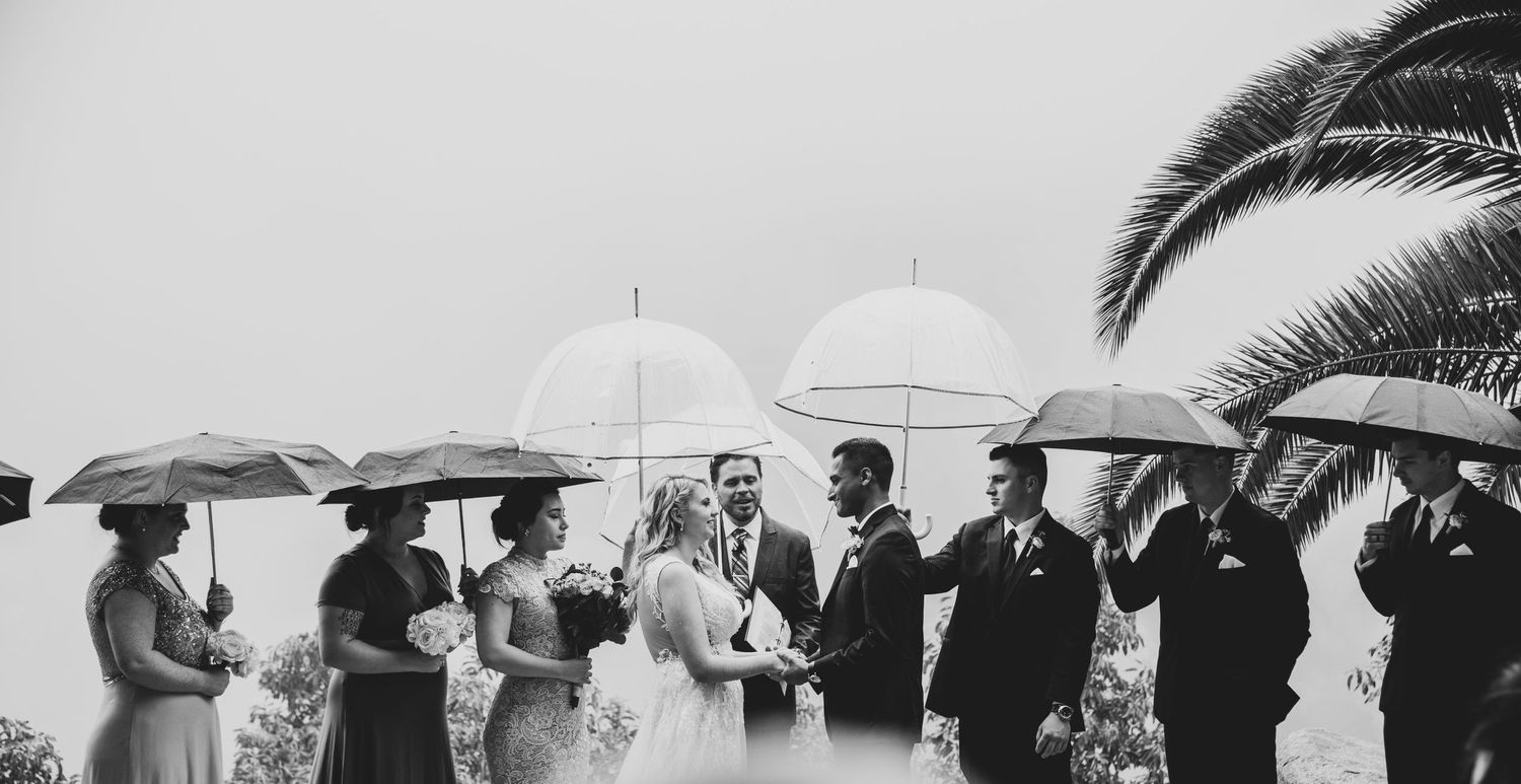 A black and white wedding ceremony captured in the rain with attendees holding white umbrellas in an outdoor setting.