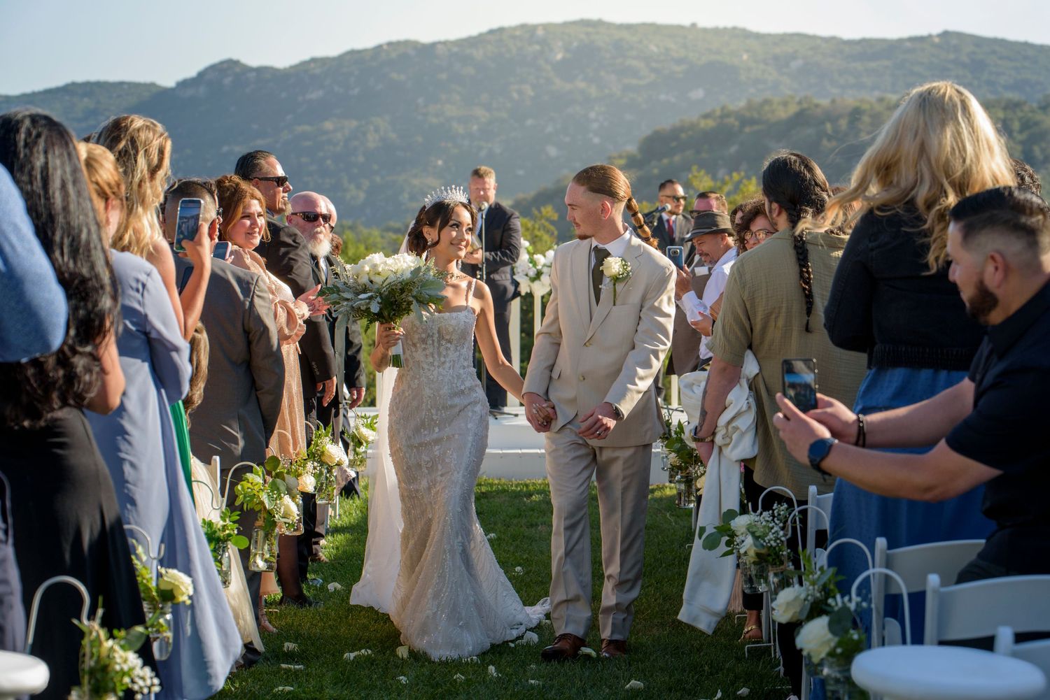 A scenic mountain wedding ceremony takes place outdoors with guests gathered around a decorated aisle at sunset.