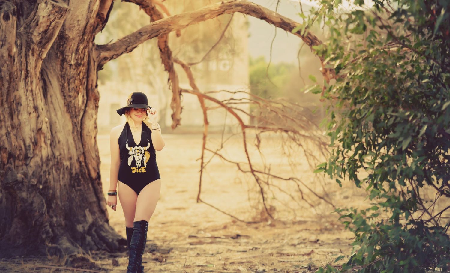 Someone in a black swimsuit and boots walks along a sun-dappled path under large twisted tree branches in a dreamy setting.