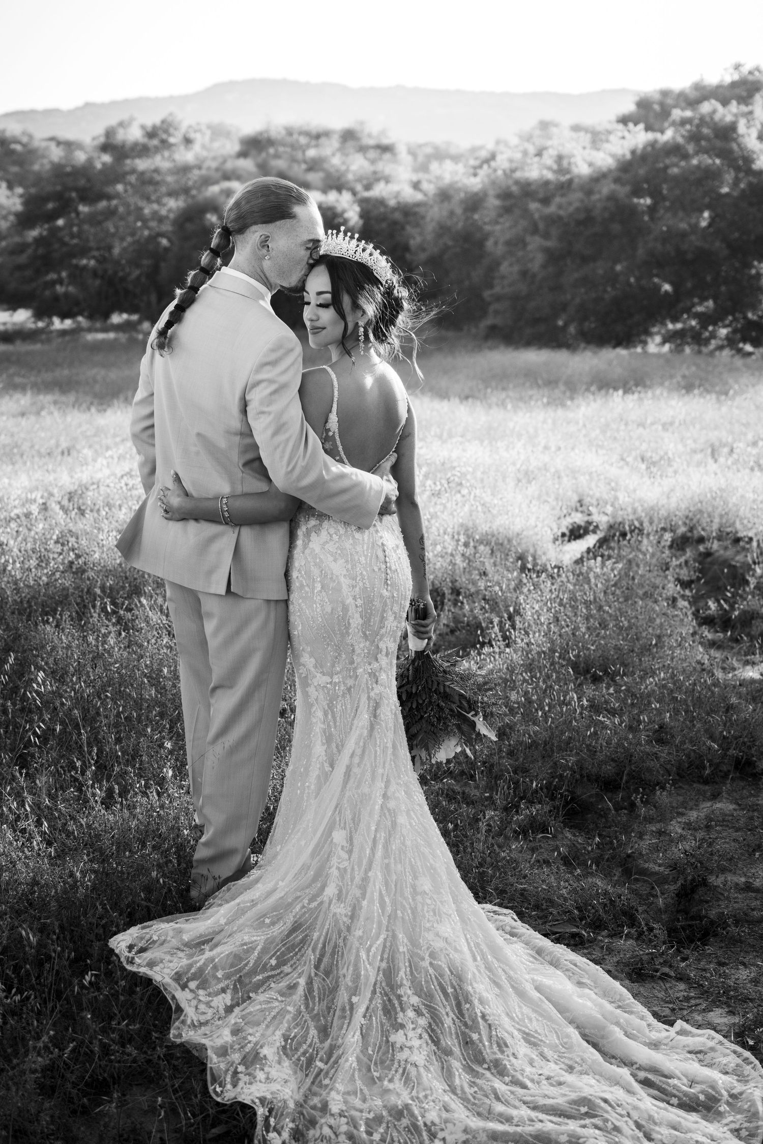 Black and white wedding photograph showing couple embracing outdoors with bride wearing stunning lace dress with long train.