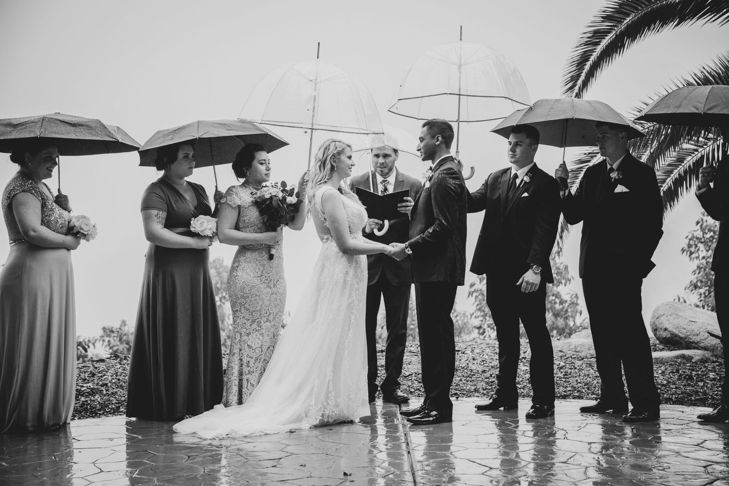 Wedding ceremony takes place outdoors in the rain with attendees holding white umbrellas during the celebration.