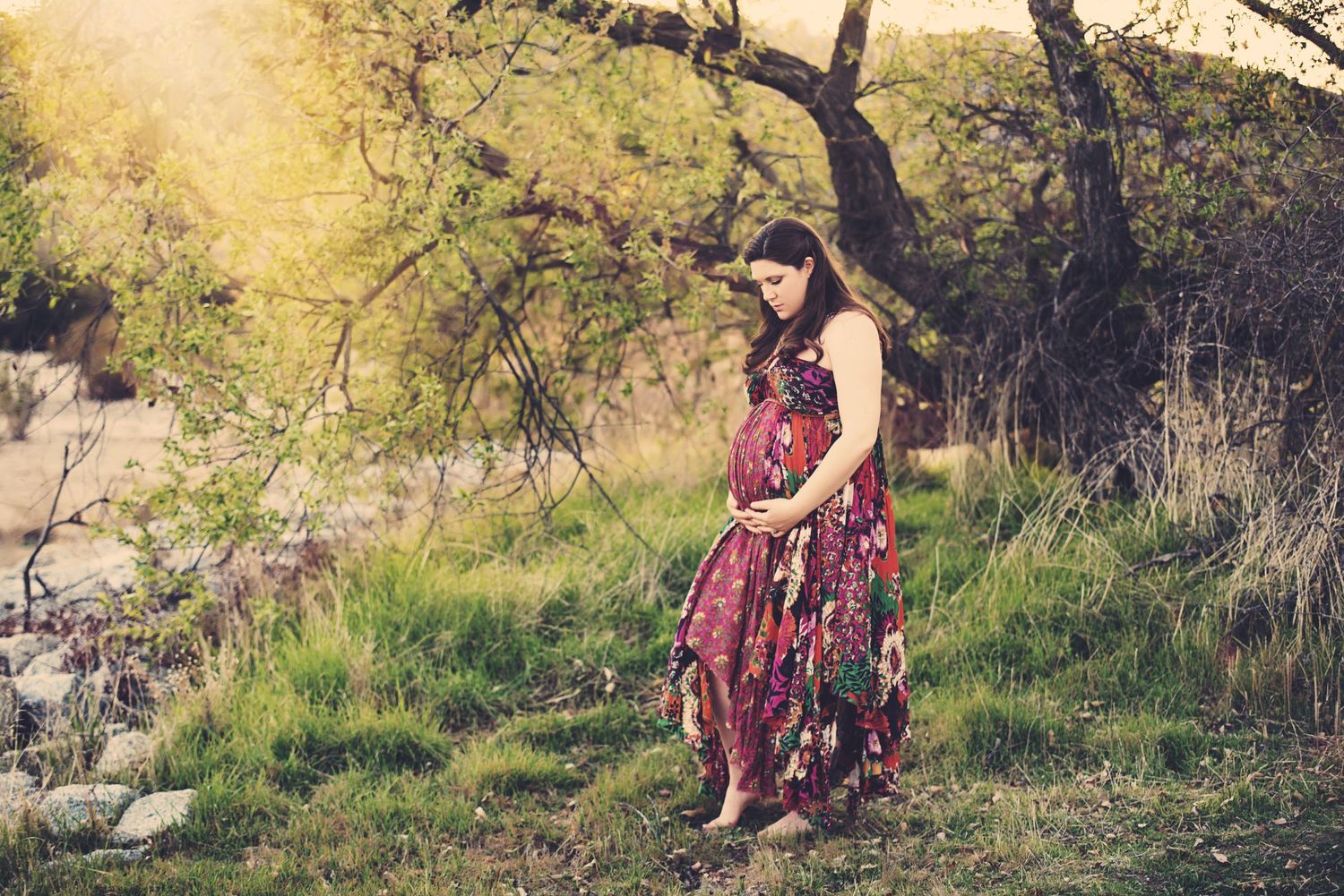 Person in a floral dress poses on a grassy hillside during golden hour among rustic tree branches.