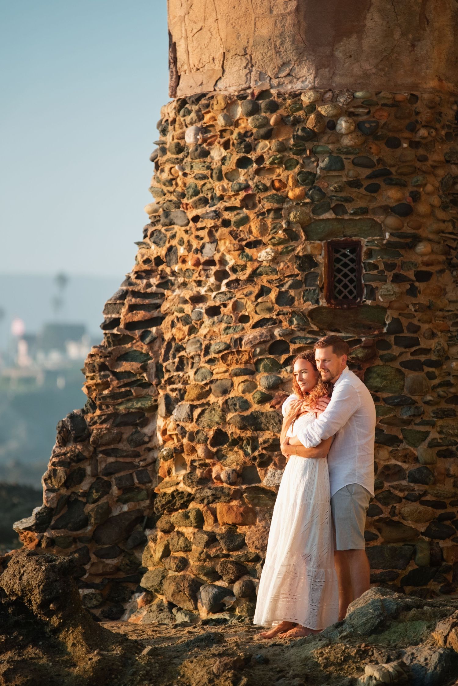 A couple embraces against a rustic stone wall during golden hour at a scenic overlook.