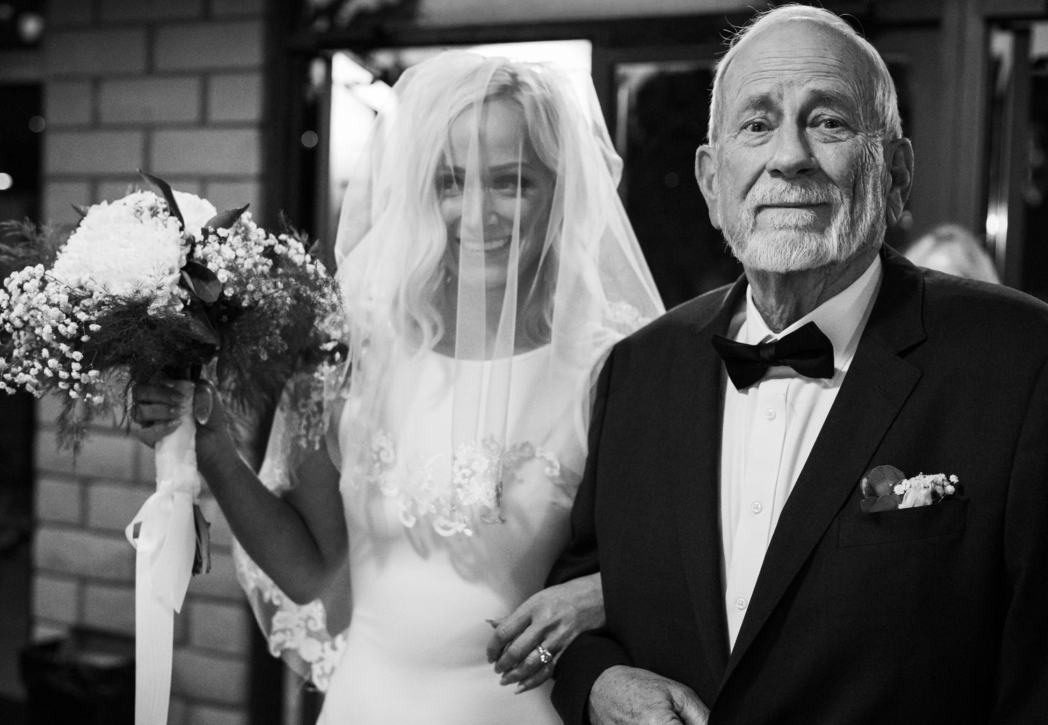 Black and white photo of a smiling bride in veil holding bouquet walking with elderly man in tuxedo at wedding.