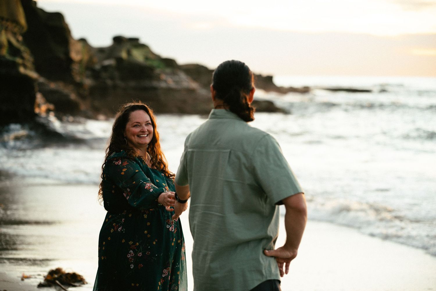 Couple holding hands on a rocky beach at sunset, woman in floral dress smiling at man in green shirt.