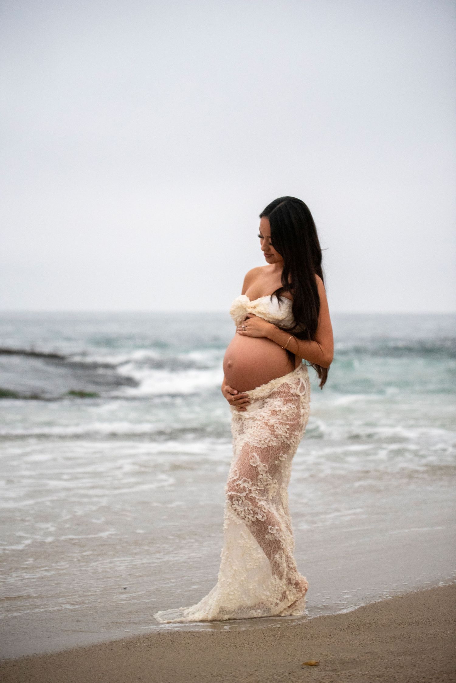 Silhouette of an expectant mother in a lace dress standing on a misty beach at sunset holding her baby bump.