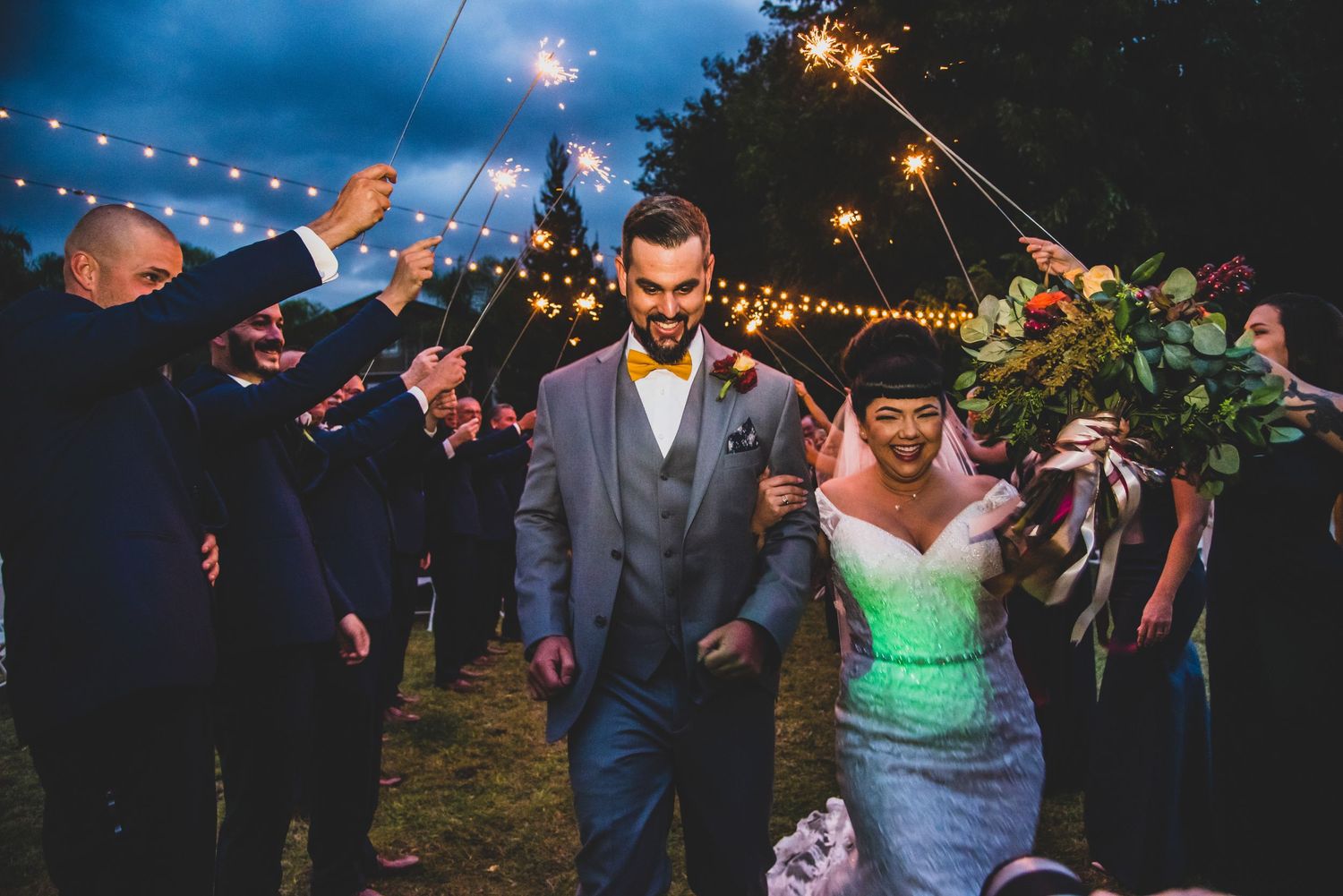 Evening outdoor wedding celebration with couple walking through sparklers under string lights in garden setting.