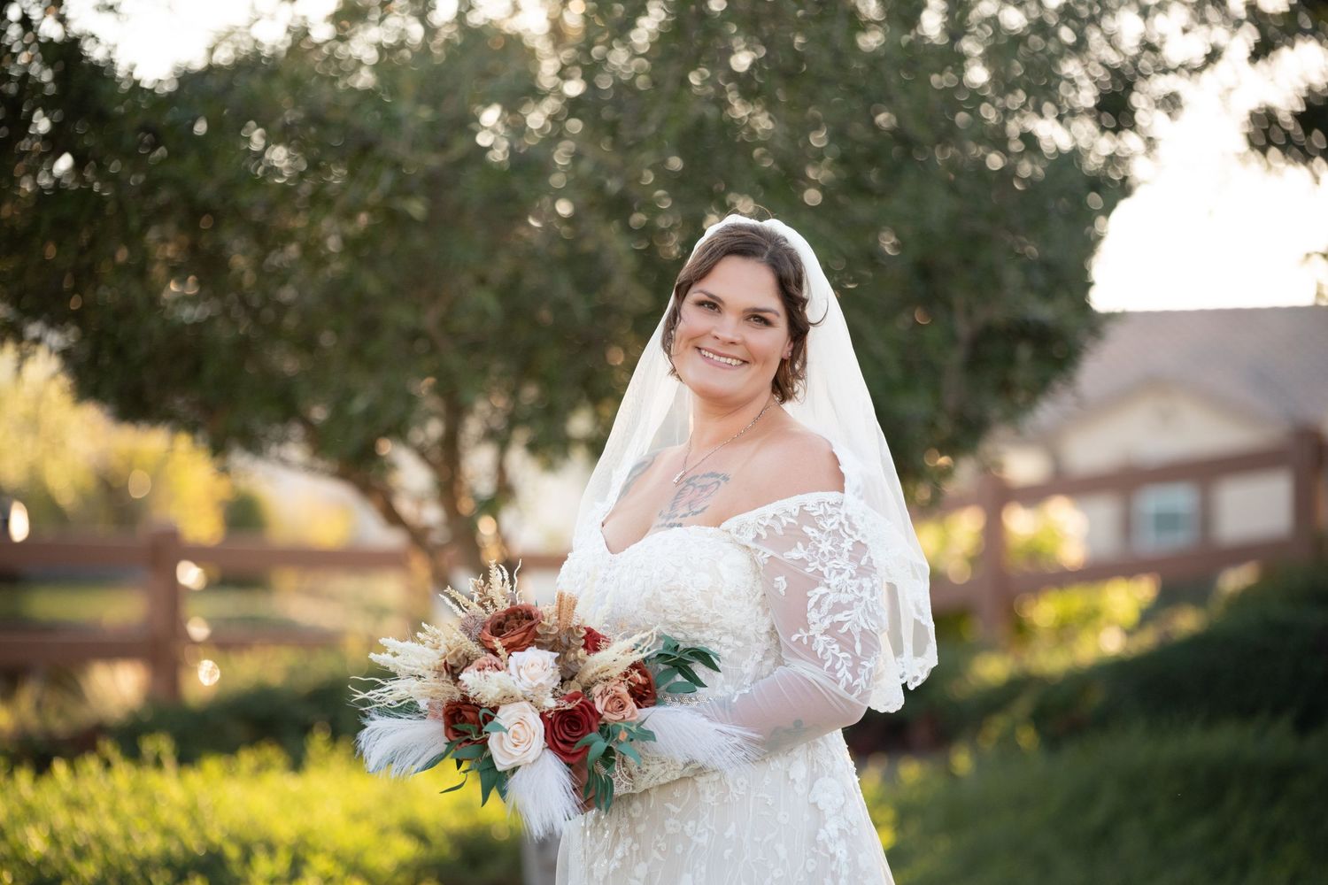 A person in a white lace wedding dress poses outdoors at sunset with a floral bouquet.