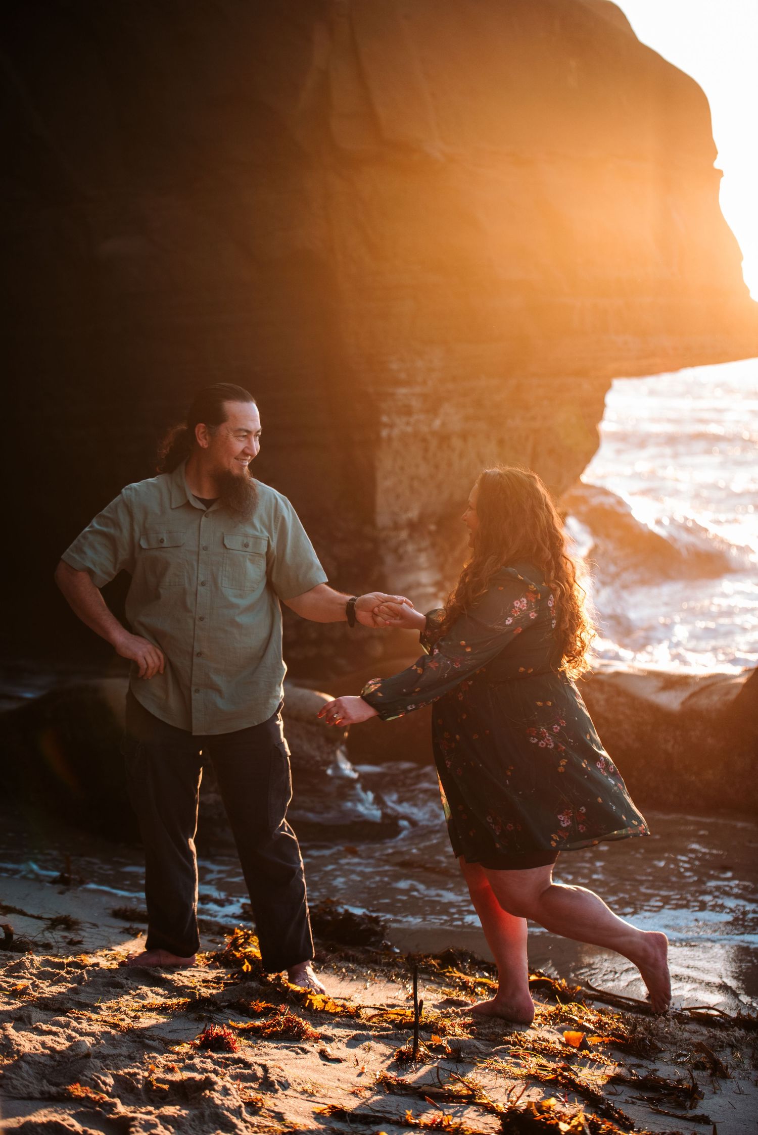 Couple holding hands and dancing on a rocky beach at golden sunset with dramatic coastal cliffs behind them.