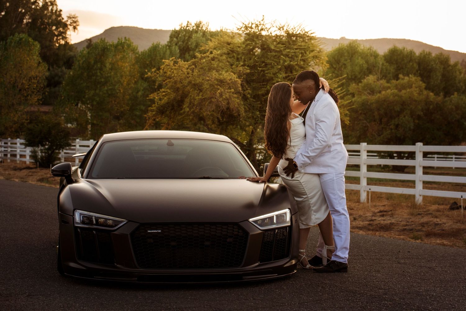 A black Audi R8 sports car parked at sunset with a white fence and mountains in the background.