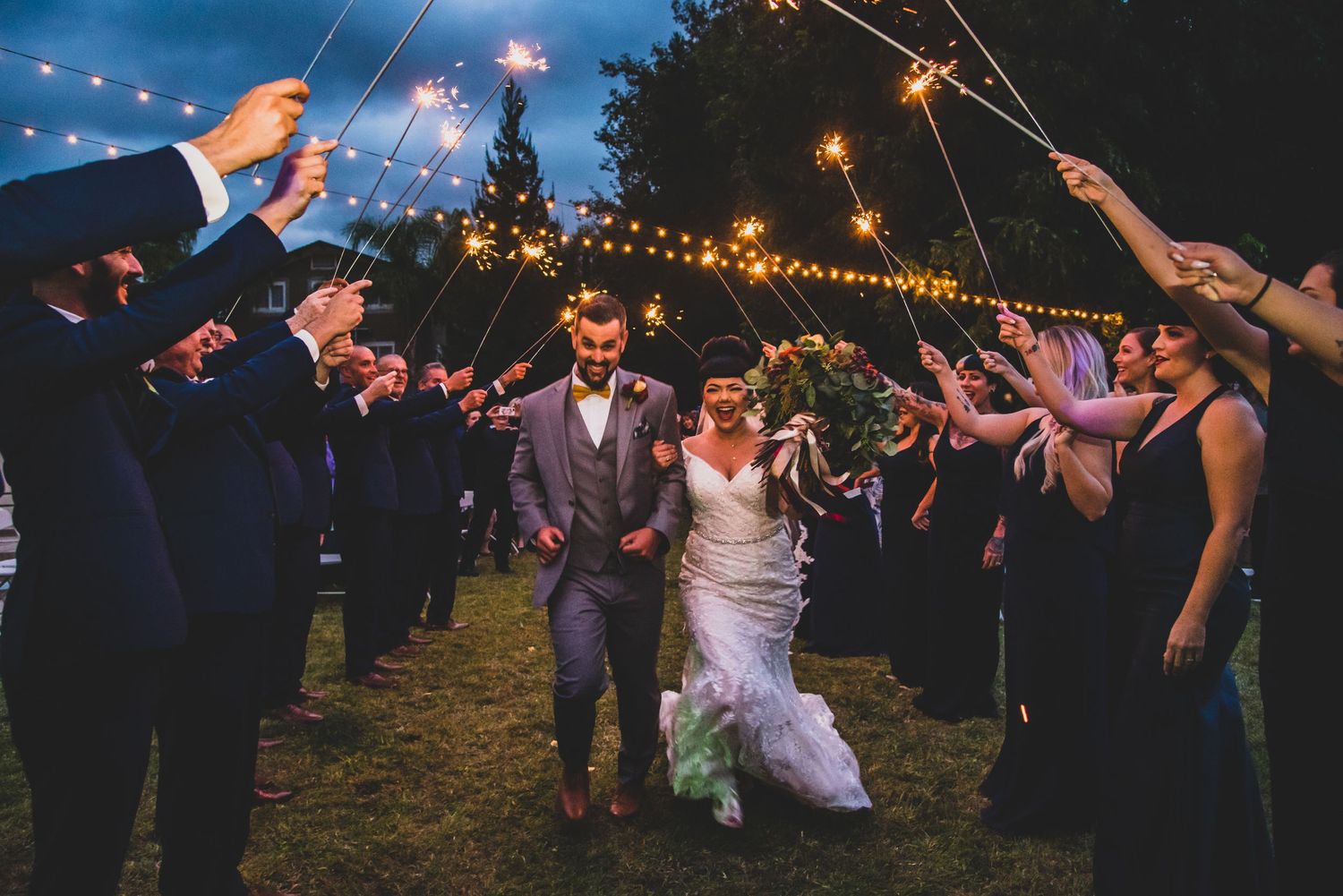Wedding guests create a sparkler tunnel exit at nighttime outdoor reception as couple walks through glowing pathway.