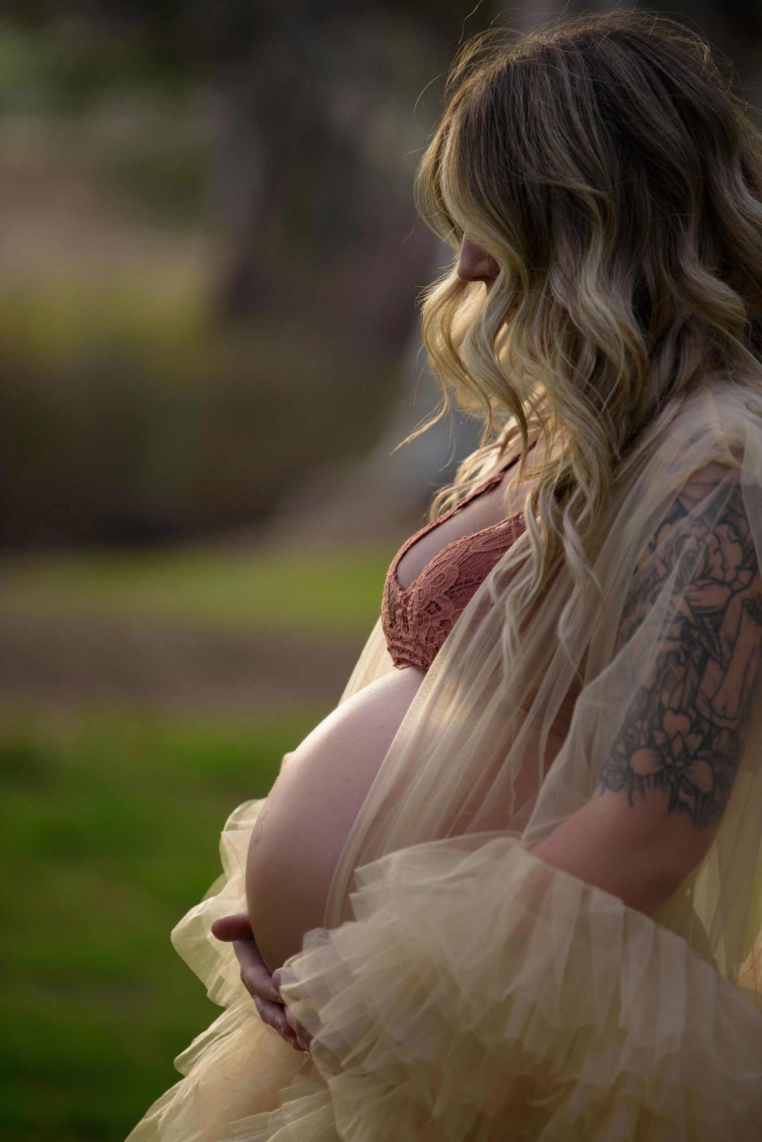 A pregnant woman in a pink lace gown with flowing blonde waves and visible tattoos poses in a dreamy outdoor setting.