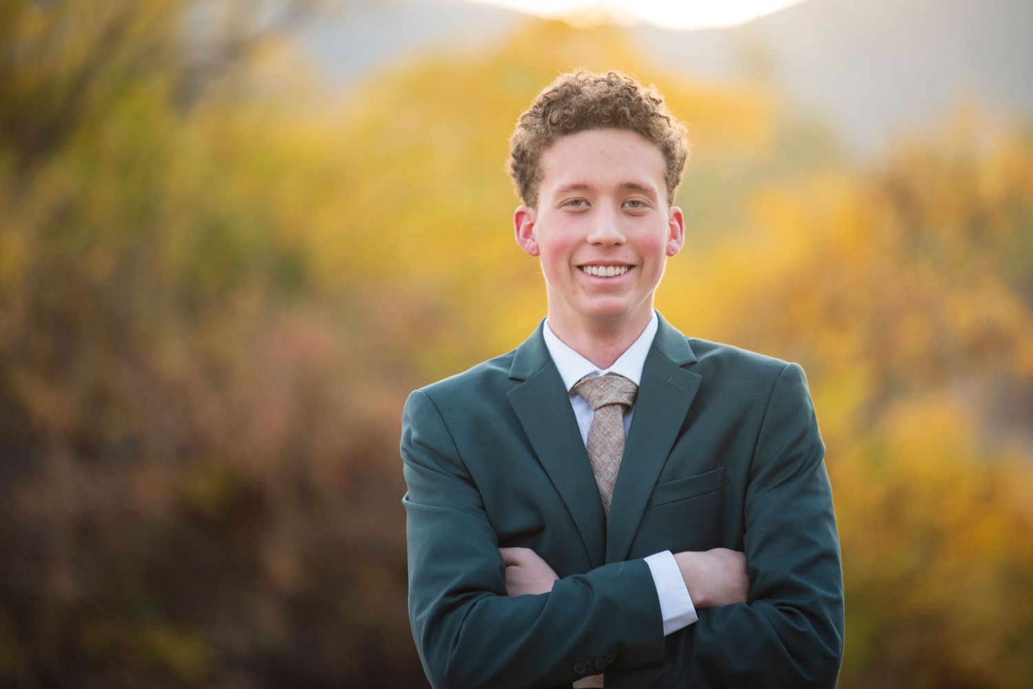 A young man in a green suit coat smiles while standing outdoors with autumn foliage in the background.
