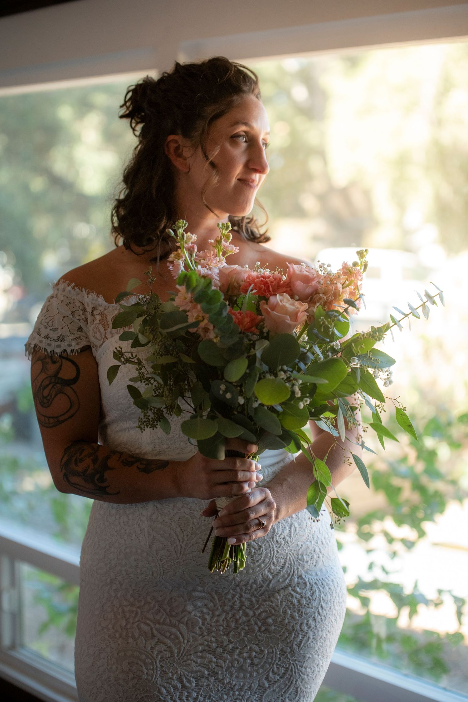 A bride in a fitted lace gown holds a beautiful pastel bouquet with pink flowers and eucalyptus by a sunlit window.