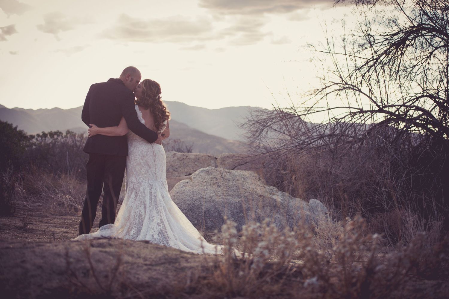 A romantic wedding couple embraces while overlooking a scenic mountain landscape at sunset.