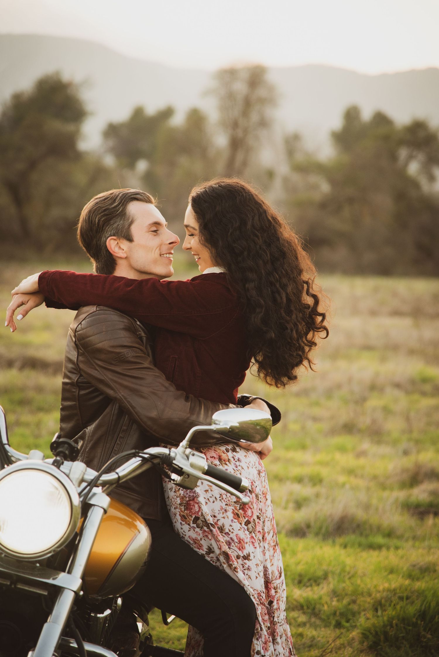 Romantic couple embraces on vintage motorcycle in sunny meadow with mountains in background.
