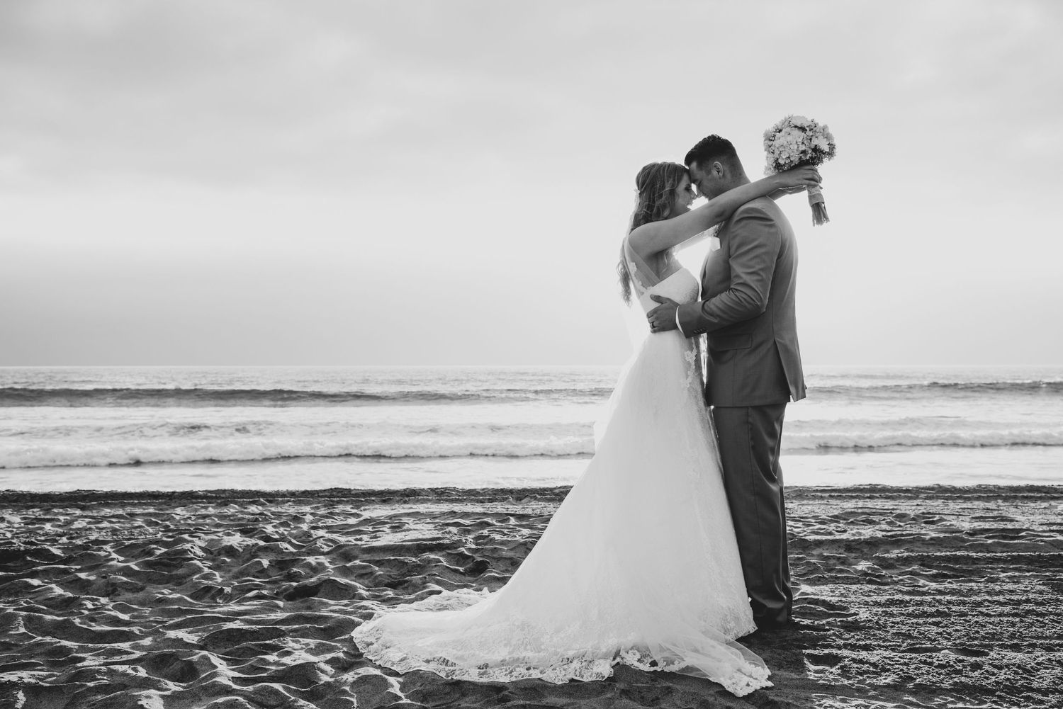 A romantic black and white beach wedding photo of a couple embracing on the sand at sunset.
