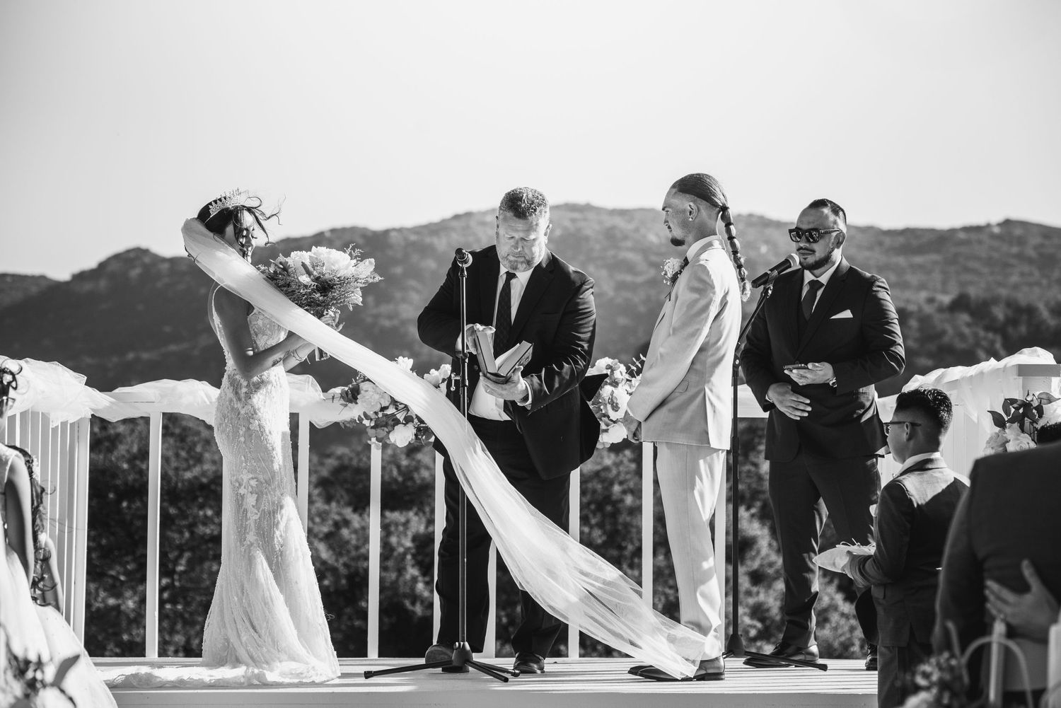 A black and white wedding ceremony photograph shows a bride in a flowing veil at an outdoor mountain venue.