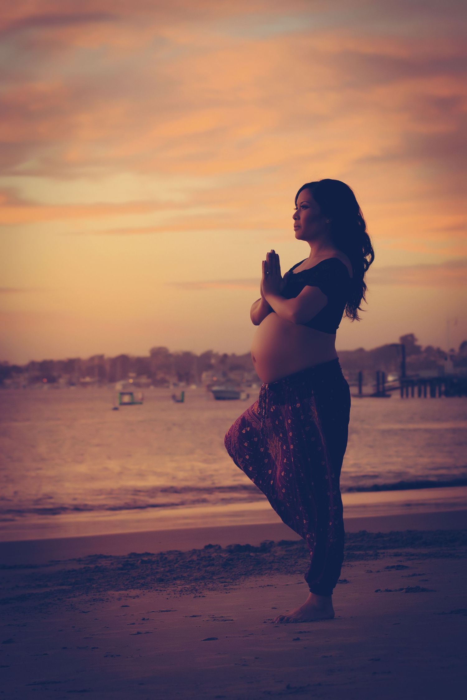 A pregnant silhouette practices yoga on a beach at sunset with boats and docks visible in the background.