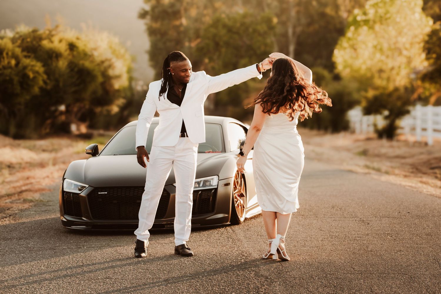 A couple in white outfits dance joyfully on a road with a black luxury car in the background during golden hour.