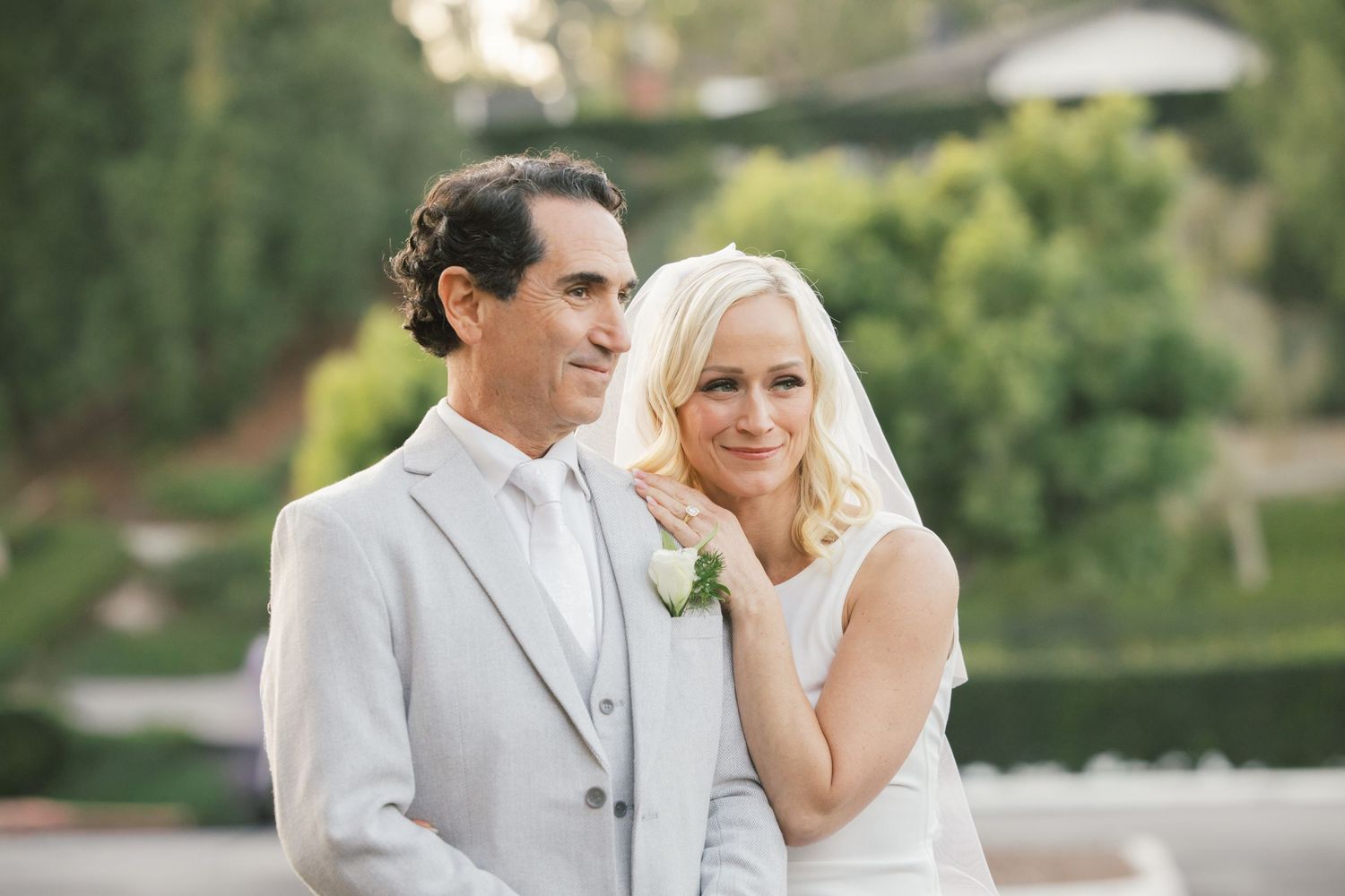 Bride and groom pose together outdoors, she in white dress with bouquet, he in light gray suit with boutonniere.
