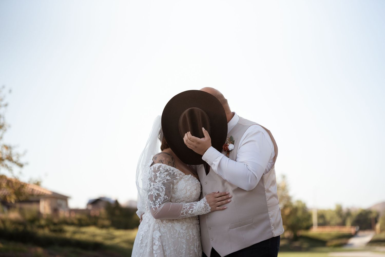 Bride and groom share a romantic kiss hidden behind a brown hat outdoors on their wedding day.