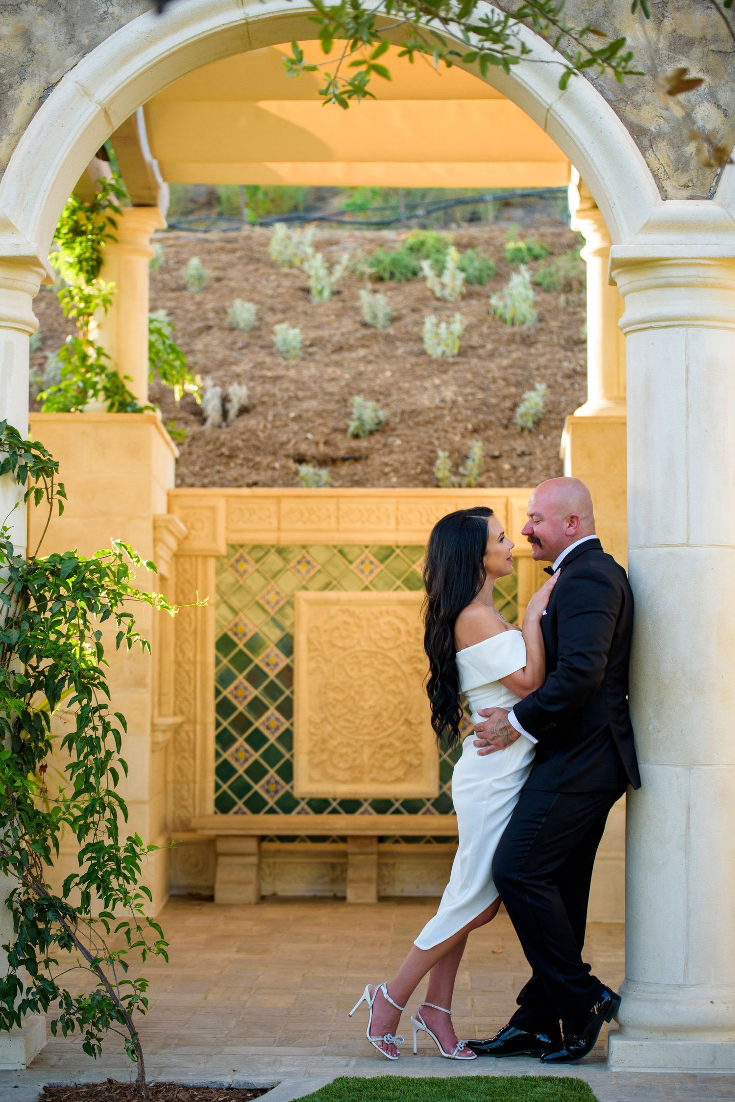 A couple embraces beneath an elegant archway decorated with climbing vines in a romantic garden setting.